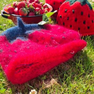 En smilende kvinde i en rød jordbærhue, ligger i græsset med en rød skål fuld af jordbær foran sig.