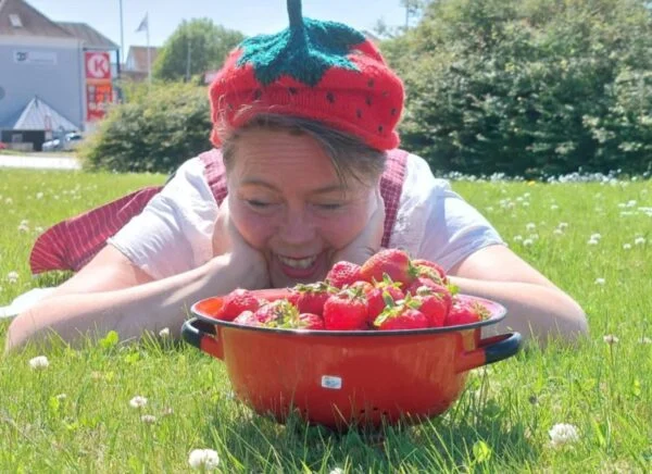 To håndstrikkede jordbærhuer i rød og grøn, omgivet af en kurv med friske jordbær, placeret på en grøn græsplæne i sollys.