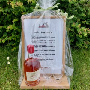 En gavekurv til snedkeren, pakket i cellofan, indeholdende en flaske mjød, to glas og et indrammet digt, alt på græsset.