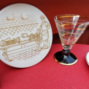 En hvid B&G platte med gylden illustration af en bager ved en maskine, ved siden af et mjødglas.