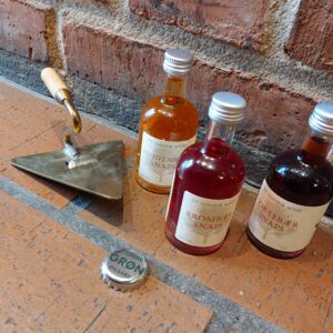 En trekantet messing øl-oplukker formet som murske, ved siden af en Tuborg Grøn kapsel og tre små flasker snaps, på mursten.