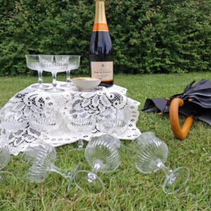 Otte antikke krystal champagneskåle og en flaske champagne på et hvidt blondedug på græsset. En paraply ligger ved siden af.