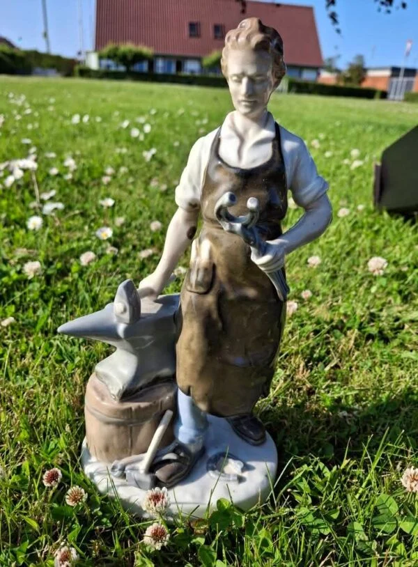 Detaljeret forside af porcelænsfiguren "Smeden", der viser figuren med forklæde og hammer ved en ambolt i græsset.