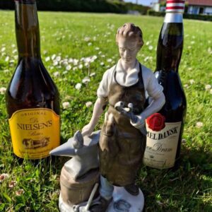 En porcelænsfigur af en beslagsmed, stående mellem to flasker spiritus, Gammel Dansk og Dr. Nielsens Bitter, på en græsplæne.