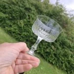 En hånd holder en klar champagneskål skråt, så kummens strålemønster og den korte stilk kan ses mod en grøn hæk.