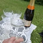 En hånd holder en bred champagneskål ved foden; flere glas, en flaske og en hvid blondedug ses bagved.
