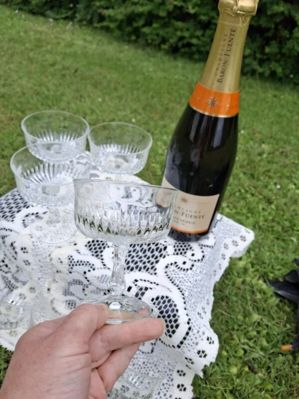 En hånd holder en bred champagneskål ved foden; flere glas, en flaske og en hvid blondedug ses bagved.