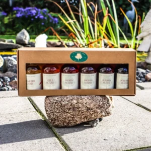 En smagsæske med seks små flasker mjød (Brombær, Æble, Hyben, Aronia, Blåbær, Solbær), præsenteret i en papæske ovenpå en stenblok udendørs.