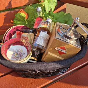 En gavekurv indeholdende en vintage kaffemølle, to pink kopper, to små snapseflasker og grønne blade, udendørs.