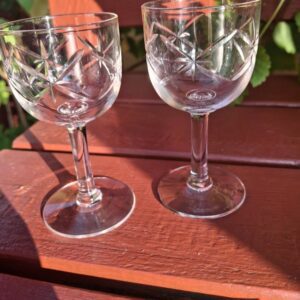 To faset slebne vintage glas på fod, med fine mønstre, placeret på en træbænk i sollys med grønne blade i baggrunden.