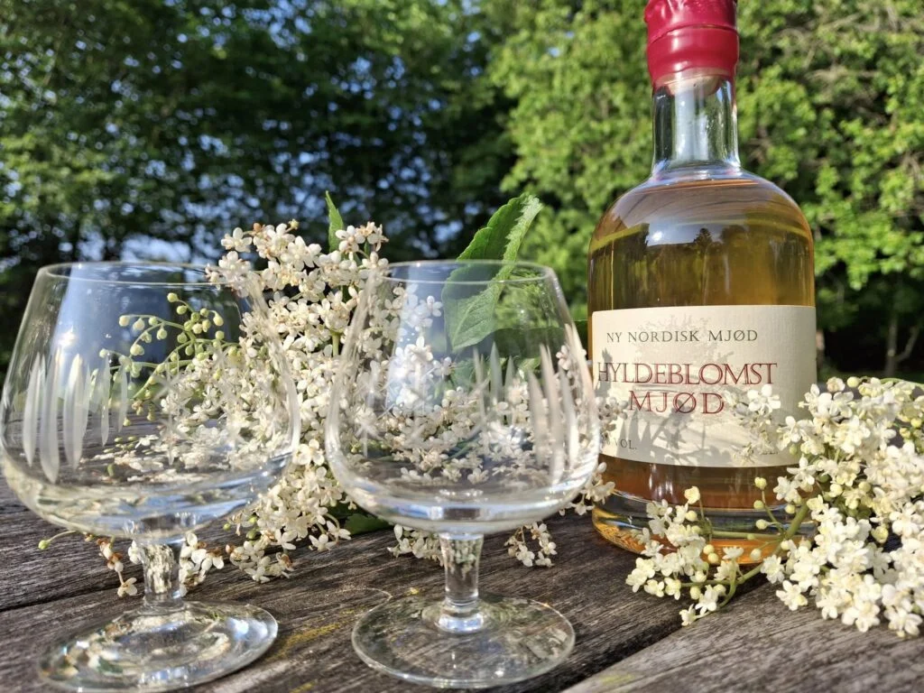 En flaske Hyldeblomst Mjød og to krystalglas, omgivet af hyldeblomster, placeret på et rustikt træbord i grønne omgivelser i sollys.