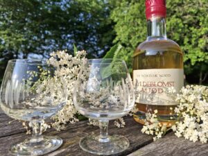 En flaske Hyldeblomst Mjød og to krystalglas, omgivet af hyldeblomster, placeret på et rustikt træbord i grønne omgivelser i sollys.