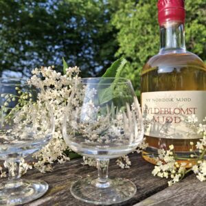 En flaske Hyldeblomst Mjød og to krystalglas, omgivet af hyldeblomster, placeret på et rustikt træbord i grønne omgivelser i sollys.