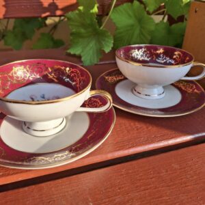 To mokkakopper med underkopper i lyserødt og guld, med blomstermønster indeni, placeret på en træbænk i sollys.