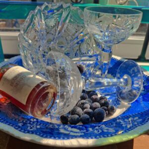 En krystalskål fyldt med frosne blåbær, liggende på en blå og hvid dekoreret platte. Et andet krystalglas og mjødflaske i baggrund.