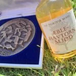 Et nærbillede af en Islandsk medalje fra 1980 i en blå gaveæske, ved siden af en lille flaske Aeblenavtorn Mjød i græsset.