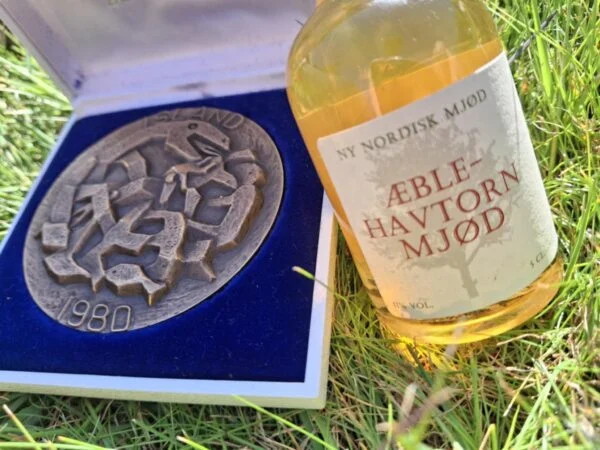 Et nærbillede af en Islandsk medalje fra 1980 i en blå gaveæske, ved siden af en lille flaske Aeblenavtorn Mjød i græsset.