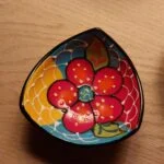 Nærbillede af en farverig, håndmalet tapasskål med et blomstermotiv i rødt, gult og blåt, på et træbord.