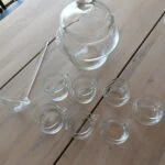 Et velkomst-drinksæt i klart glas med en bowle, seks små kopper med hanke og en øseske, arrangeret på et træbord.