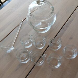 Et velkomst-drinksæt i klart glas med en bowle, seks små kopper med hanke og en øseske, arrangeret på et træbord.