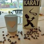 En hvid Karat kaffedåse med en sort silhuet af en kaffeserverende mand, omgivet af kaffebønner og en hvid kop på træbord.