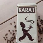 Et nærbillede af en vintage Karat kaffedåse med logoet og en kaffeserverende silhuet, omgivet af spredte kaffebønner på et træbord.