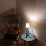 En Bombay Sapphire gin flaske lampe med hvid skærm, placeret på et lille blåt bord ved siden af en gyngestol i et hjørne af rummet.
