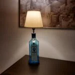En Bombay Sapphire gin flaske omdannet til en lampe med en hvid lampeskærm, står på et mørkt bord i et hyggeligt rum.