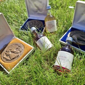Flere åbne gaveæsker med Nordatlantiske medaljer, flankeret af små flasker mjød og snaps, alt på grønt græs.