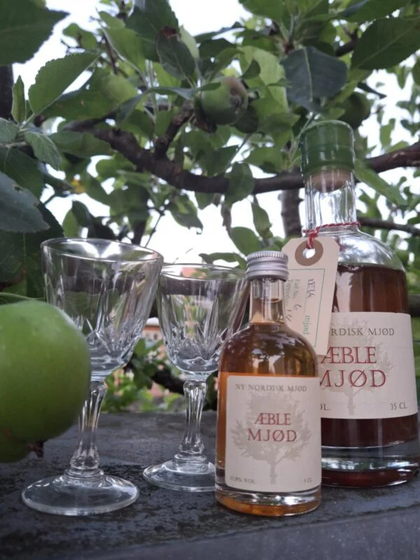 To klare glas med mønster nederst på kummen ved to flasker æblemjød under et æbletræ.