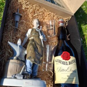 To tomme dramglas med teksten 'En lille dram', placeret i en gavekasse med halm, ved siden af en beslagsmed figur.