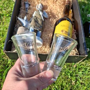 To klare dramglas med teksten 'En lille dram' i sort skrift, holdt op foran en gavekurv.