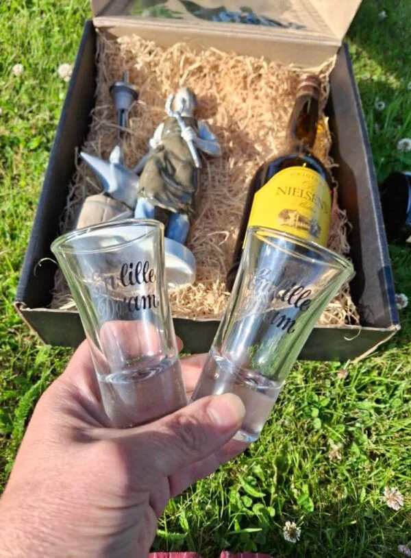 To klare dramglas med teksten 'En lille dram' i sort skrift, holdt op foran en gavekurv.