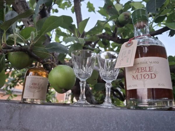 To mønstrede glas på fod mellem to flasker æblemjød med grønne æbler over.