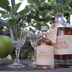 To krystalglas og to flasker Aeblen Mjød – en stor og en lille – placeret under et æbletræ med grønne æbler.