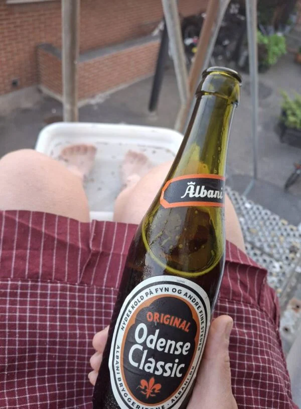 En person slapper af med fødderne i et fodbad og holder en flaske Odense Classic øl. Set fra personens perspektiv.