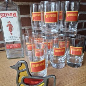 En tom Beefeater London Dry Gin flaske, stablede gin glas med rødt logo og en fugleformet oplukker, alt på et træbord.