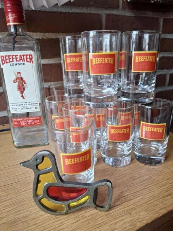 En tom Beefeater London Dry Gin flaske, stablede gin glas med rødt logo og en fugleformet oplukker, alt på et træbord.