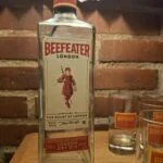 Den ikoniske Beefeater London Dry Gin flaske flankeret af matchende gin glas med rødt logo på et træbord.