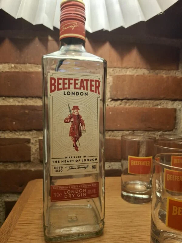 Den ikoniske Beefeater London Dry Gin flaske flankeret af matchende gin glas med rødt logo på et træbord.