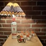 Beefeater gin flaske omdannet til bordlampe med plisseret skærm, omgivet af gin glas og fugle-oplukker. Står på træbord foran murstensvæg.