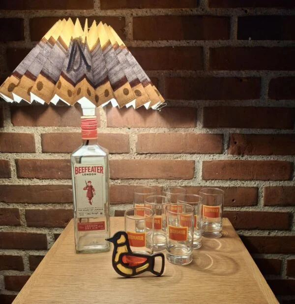 Beefeater gin flaske omdannet til bordlampe med plisseret skærm, omgivet af gin glas og fugle-oplukker. Står på træbord foran murstensvæg.