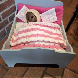 En mørkhudet dukke iført hæklet hue i en lyseblå vintage dukkeseng med lyserødt og hvidt sengetøj.