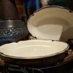 To hvide, ovale serveringsskåle med guldkant, tilhørende et vintage stel, placeret på et træunderlag.