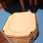En stabel af kvadratiske, hvide frokosttallerkener med guldkant, tilhørende et vintage stel, placeret på en blå overflade.