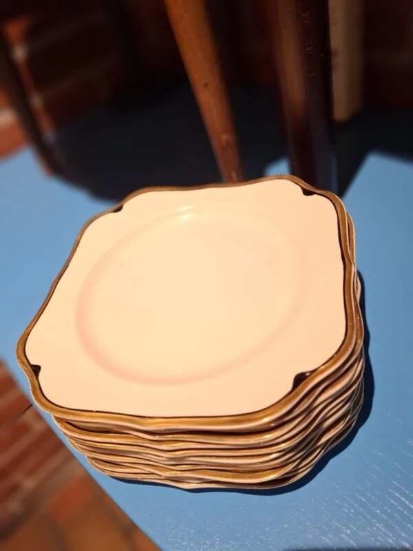 En stabel af kvadratiske, hvide frokosttallerkener med guldkant, tilhørende et vintage stel, placeret på en blå overflade.