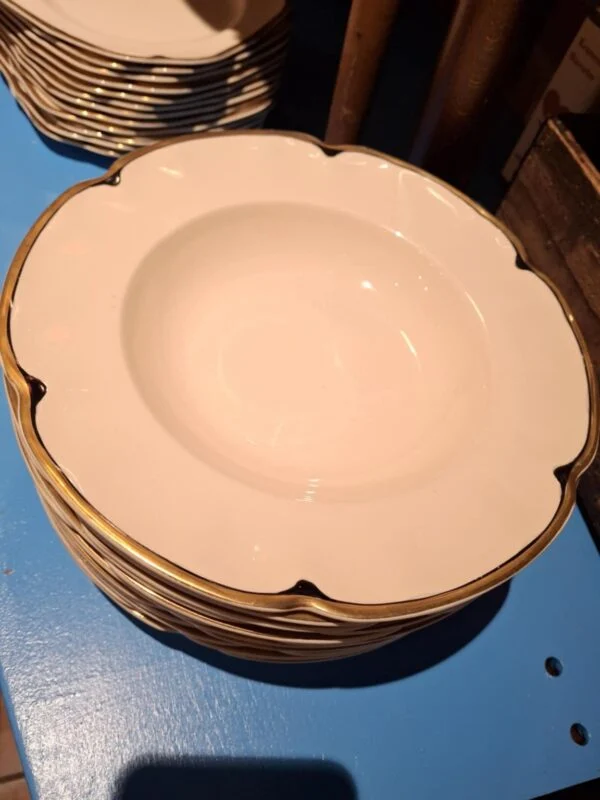 En stabel af dybe, hvide tallerkener med guldkant, tilhørende et vintage stel, placeret på en blå overflade.