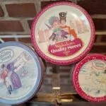Tre vintage Quality Street dåser i forskellige størrelser, den mindste rund og rød, mod en murstensvæg.