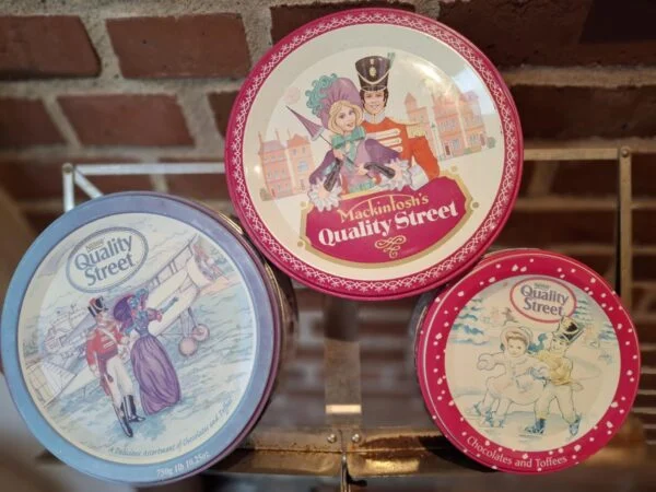 Tre vintage Quality Street dåser i forskellige størrelser, den mindste rund og rød, mod en murstensvæg.