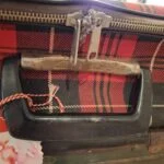 Nærbillede af håndtaget og den kraftige lynlås på en rødrutet vintage kuffert, med et lille hjertevedhæng på lynlåsen.